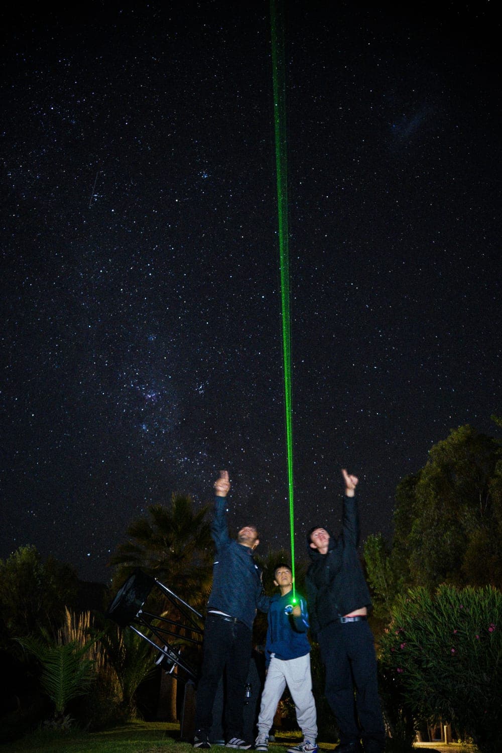Tres personas apuntan al cielo mientras un láser verde sube entre las estrellas, junto a un telescopio en El Molle.